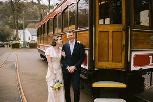 Bride and groom smiling together beside a vintage red trolley car on their wedding day.