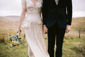 Bride in ivory gown holding sunflower bouquet walks hand-in-hand with groom in black suit across scenic highland meadow.