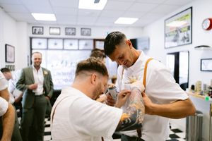 Two barbers in white shirts and suspenders work in a classic barbershop with checkered floors and framed art on walls.