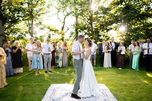 Bride and groom share first dance on outdoor rug surrounded by wedding guests under sunlit trees.