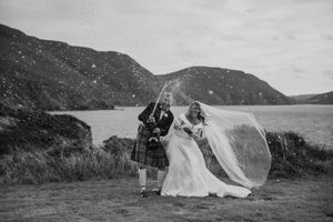 Groom in tartan kilt pops champagne beside bride in flowing veil by Scottish loch with misty hills.