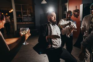 Bearded man dancing at a party with friends holding wine glasses in a dimly lit venue.