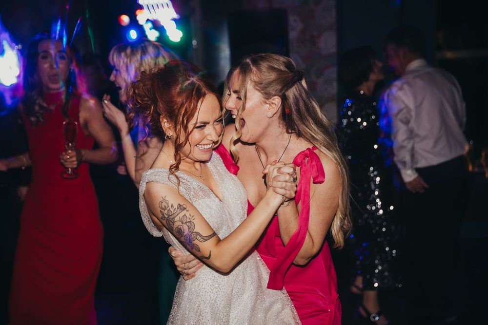 Two women dancing and embracing at a wedding reception, one in a white bridal gown and one in a red dress.