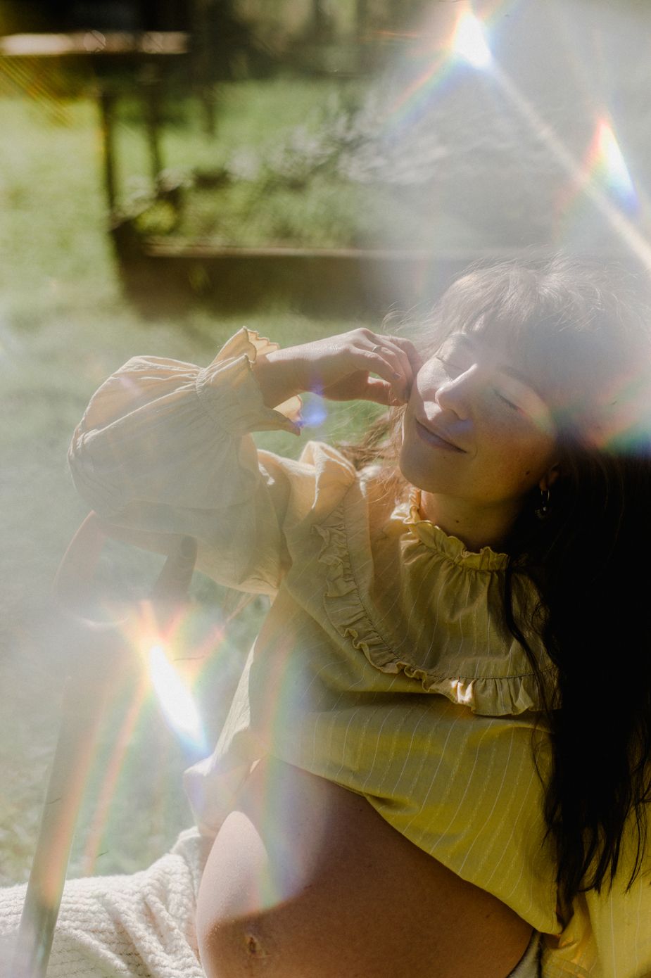 A woman with dark hair reclines outdoors in a yellow top, bathed in golden sunlight with rainbow lens flare effects.