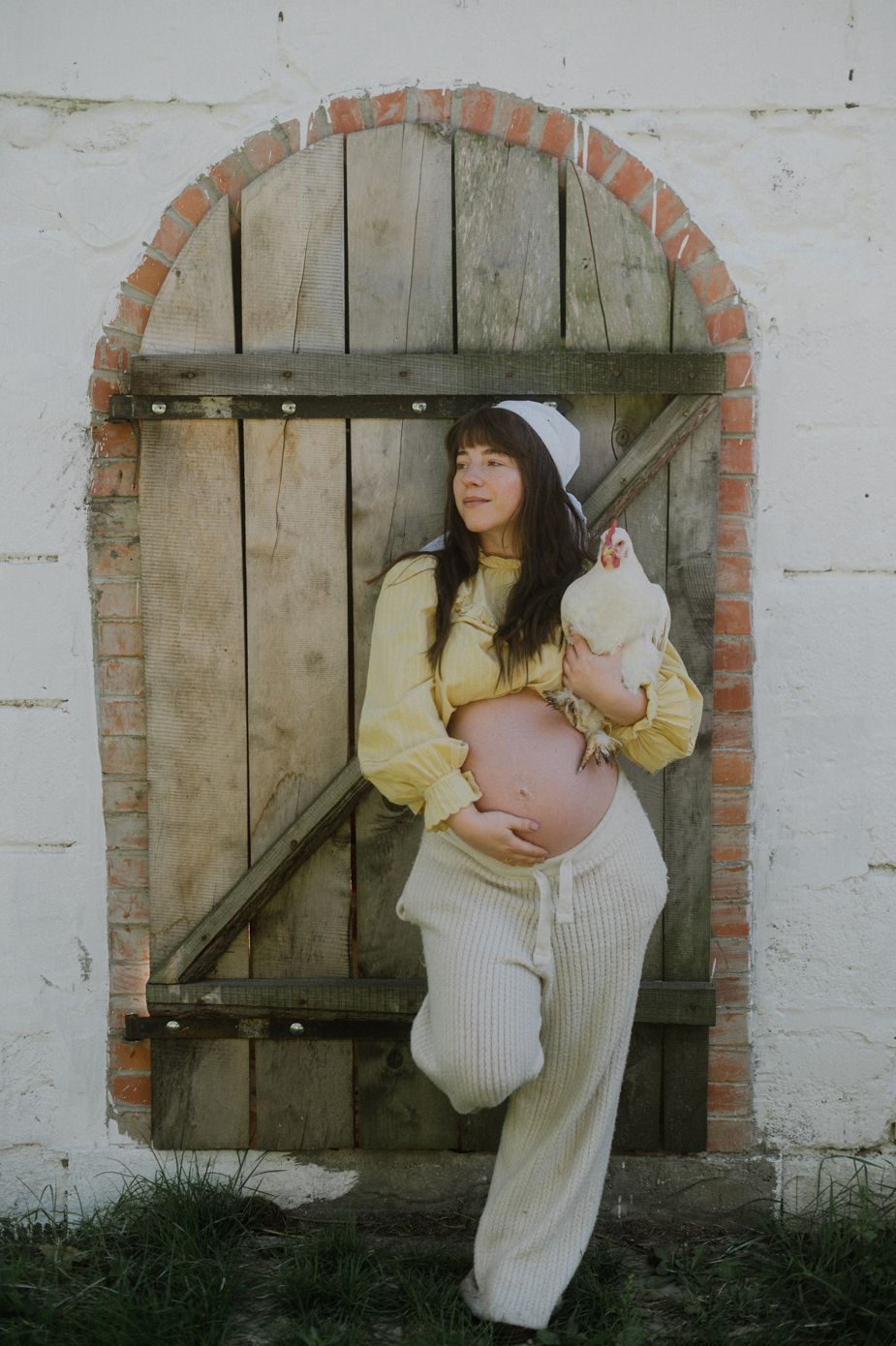 Pregnant woman in yellow top holds a white chicken, posing by a rustic arched wooden door on a farm.