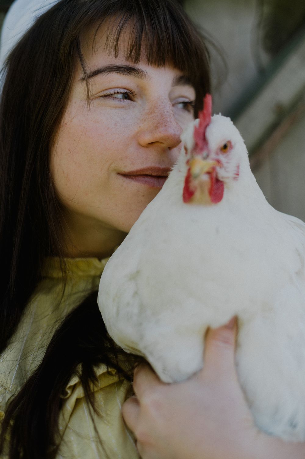 A young woman with freckles and dark hair holds a white chicken close to her face, smiling warmly outdoors.