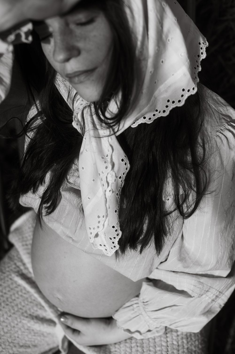 Black and white photo of a pregnant woman in a white eyelet blouse and headscarf, cradling her bare baby bump.