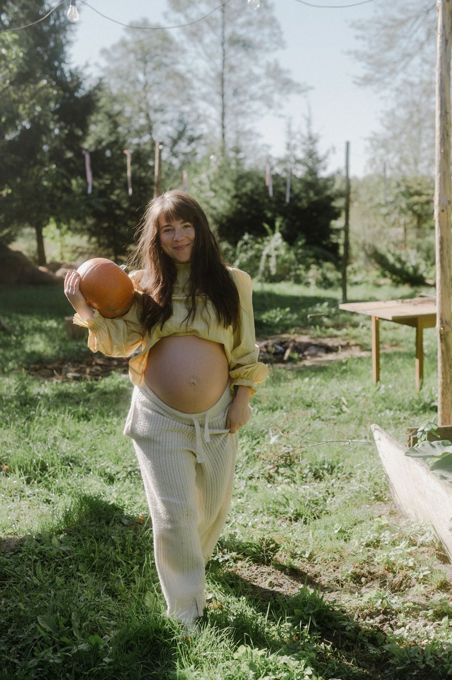 Pregnant woman in yellow top and cream pants holds a pumpkin outdoors in a sunny green garden setting.
