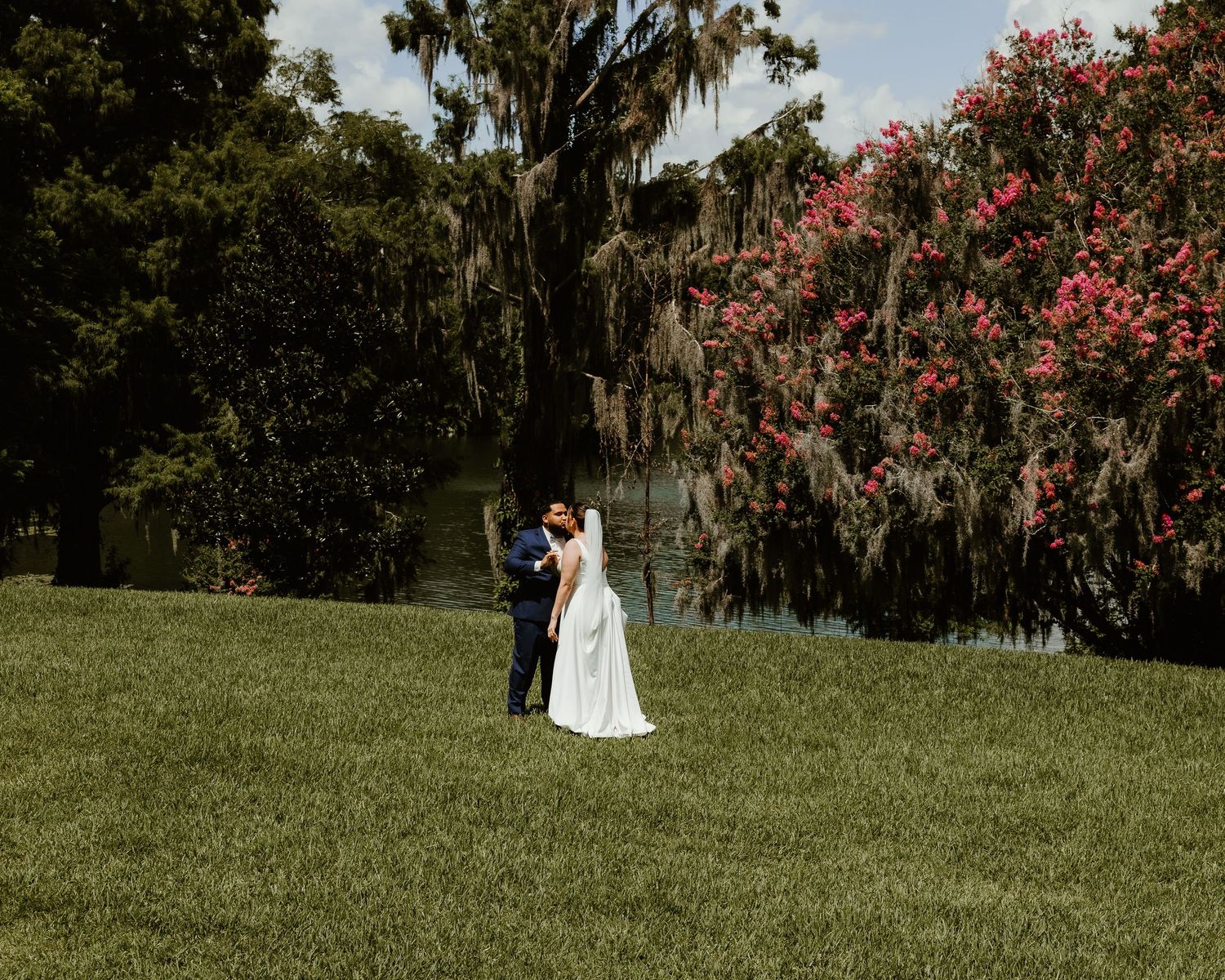 Bride and groom embrace on lush green lawn surrounded by Spanish moss trees and blooming pink flowers near a serene pond.