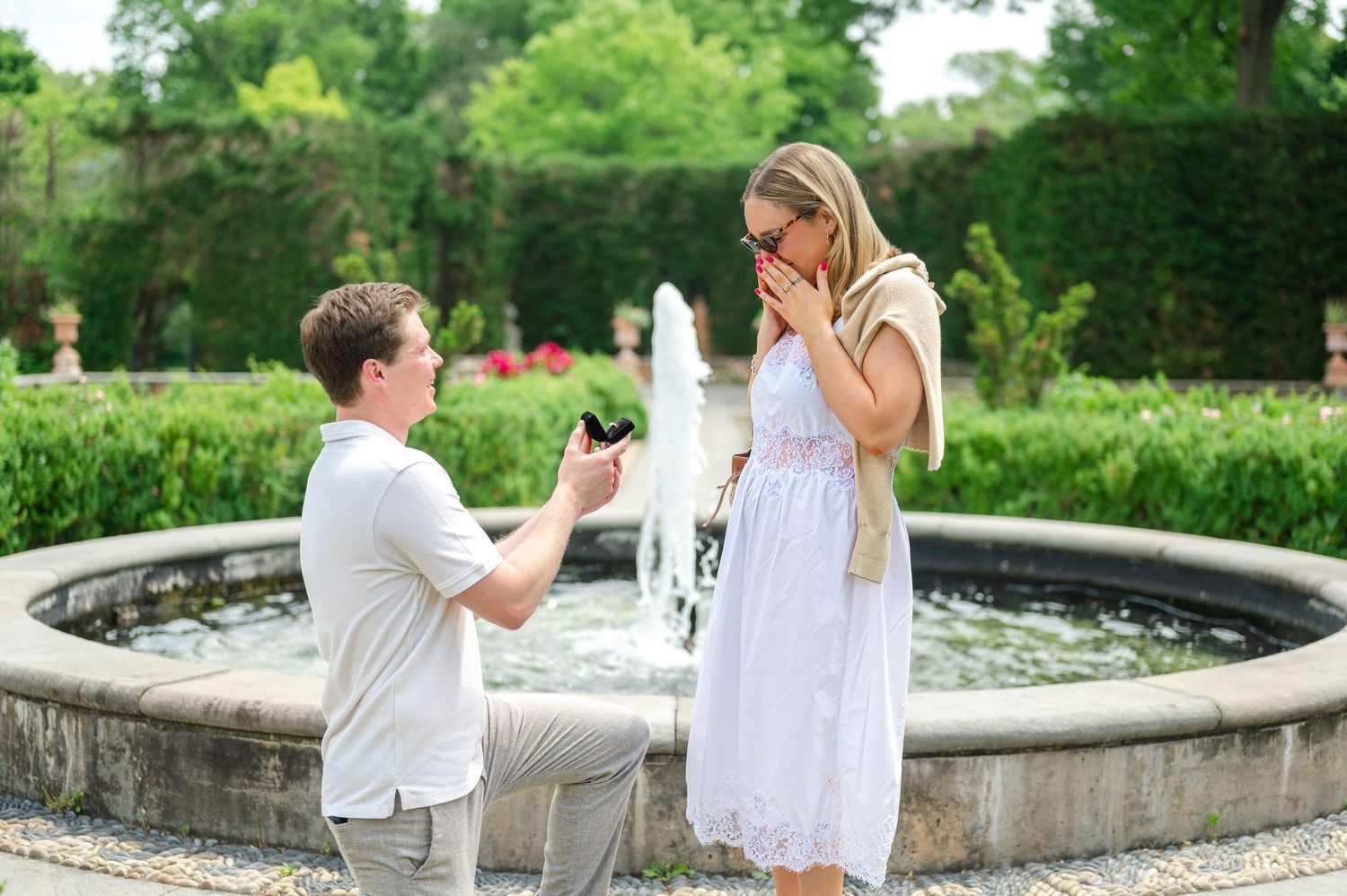 Man kneeling on one knee proposing to surprised woman outdoors in lush green garden setting.