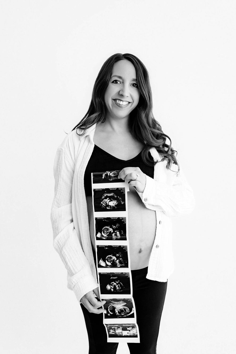 Pregnant woman smiling and holding ultrasound strip photos against her baby bump in black and white portrait.