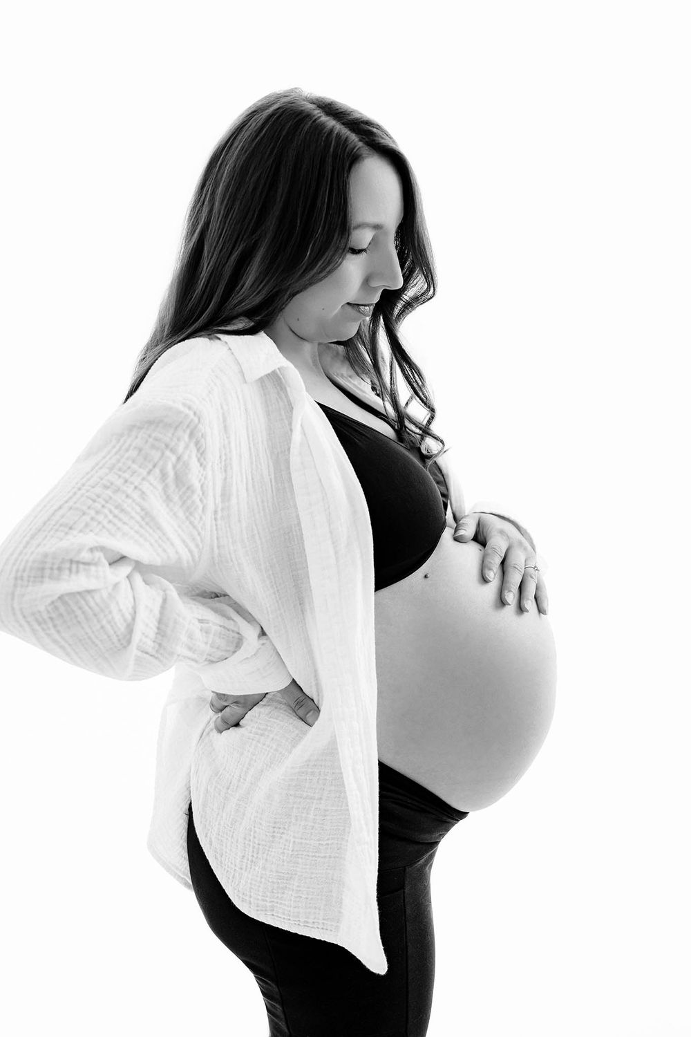 Black and white maternity photo of a pregnant woman in a white shirt cradling her baby bump against a white background.