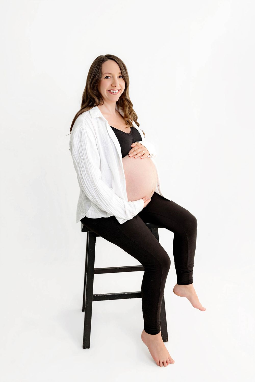 Smiling pregnant woman sitting on black stool, wearing white shirt and black leggings, cradling baby bump against white background.