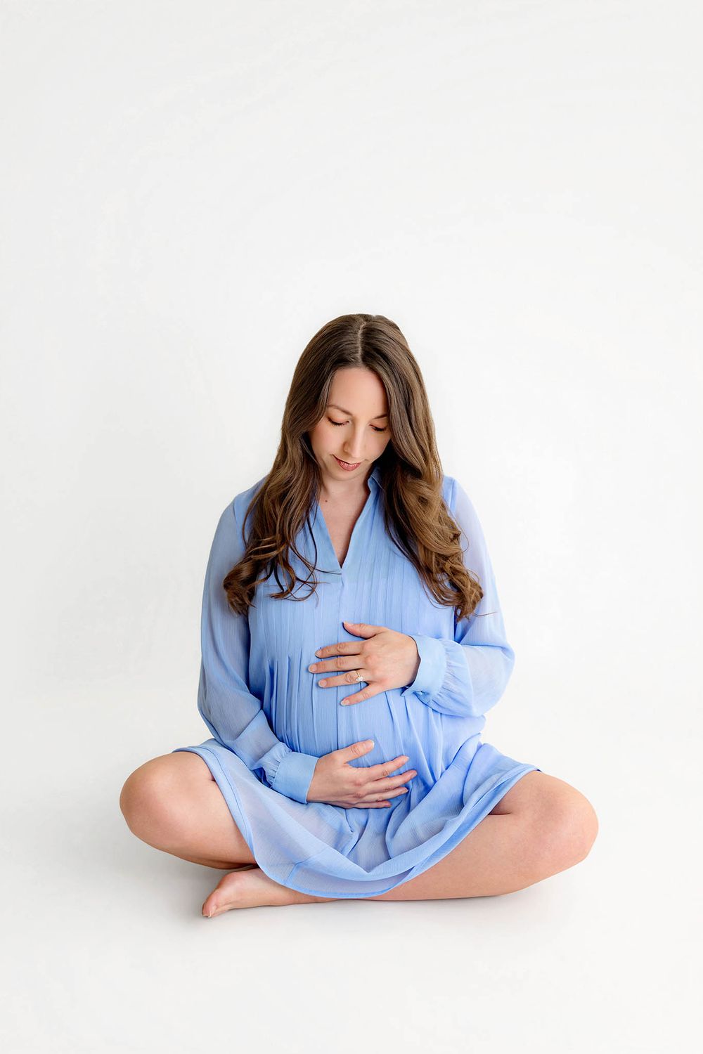 Pregnant woman in blue dress sitting cross-legged on white background, cradling her baby bump with both hands.