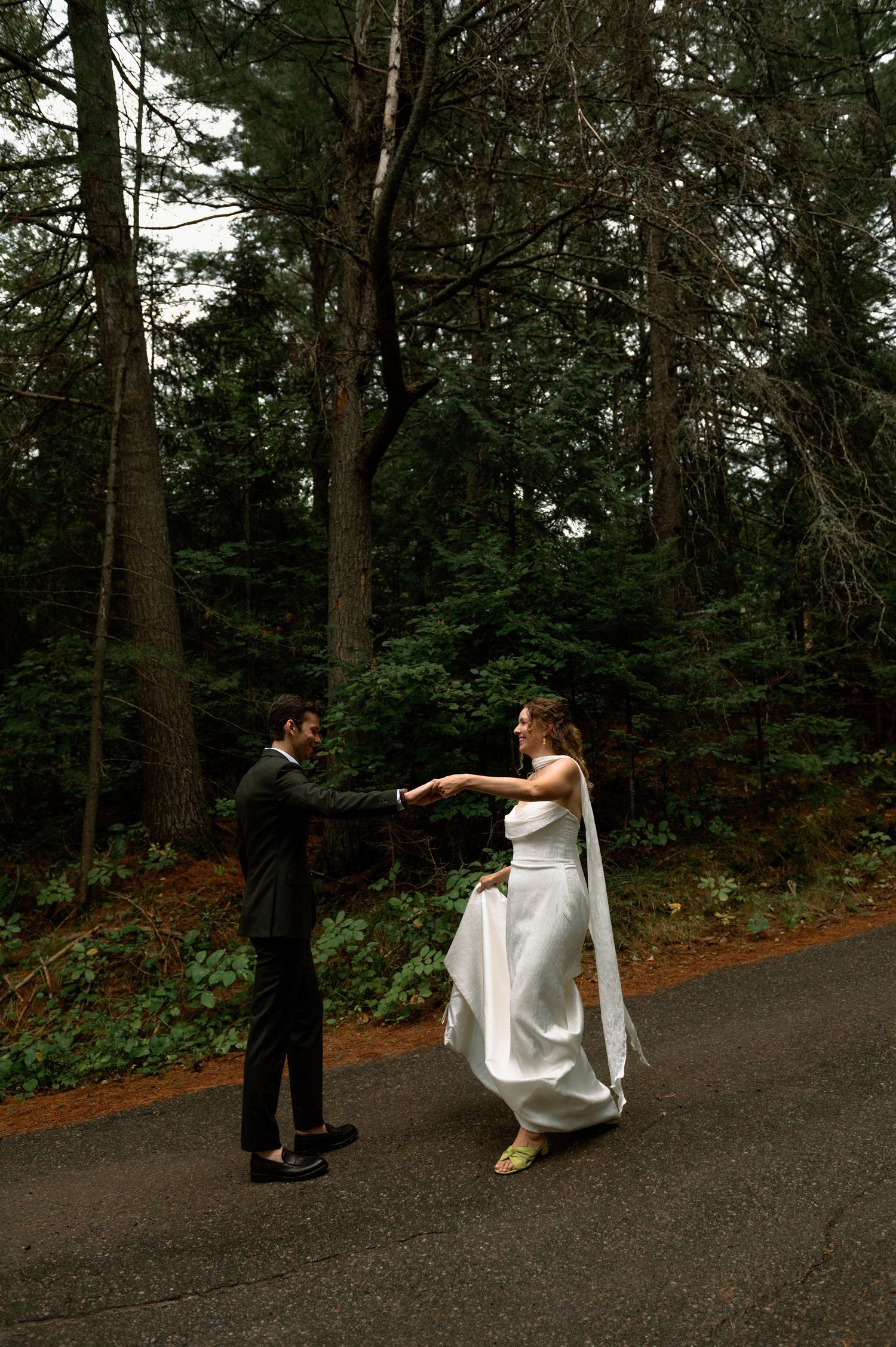 Bride in white gown and groom in black suit dance on a forest road surrounded by tall evergreen trees.