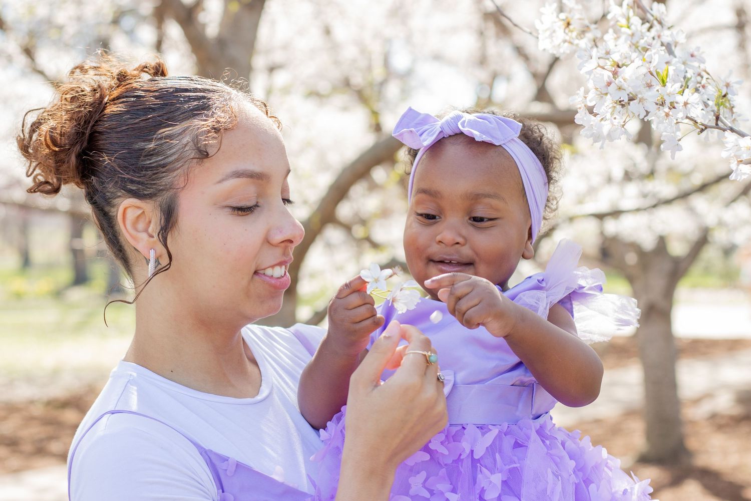 A young woman holds a baby girl in matching purple outfits among blooming white trees outdoors.