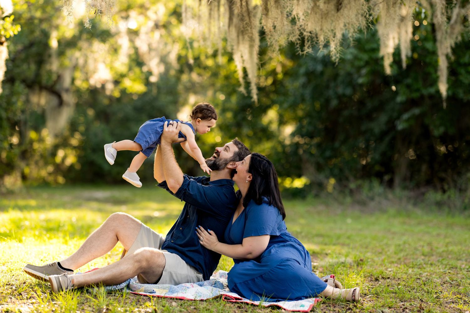 Family sitting on blanket in park, parents lifting baby girl under Spanish moss-draped trees in golden sunlight.