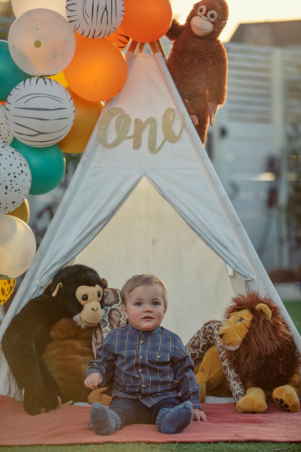 Toddler boy sits in front of a teepee with stuffed animals and colorful balloons at a first birthday party.