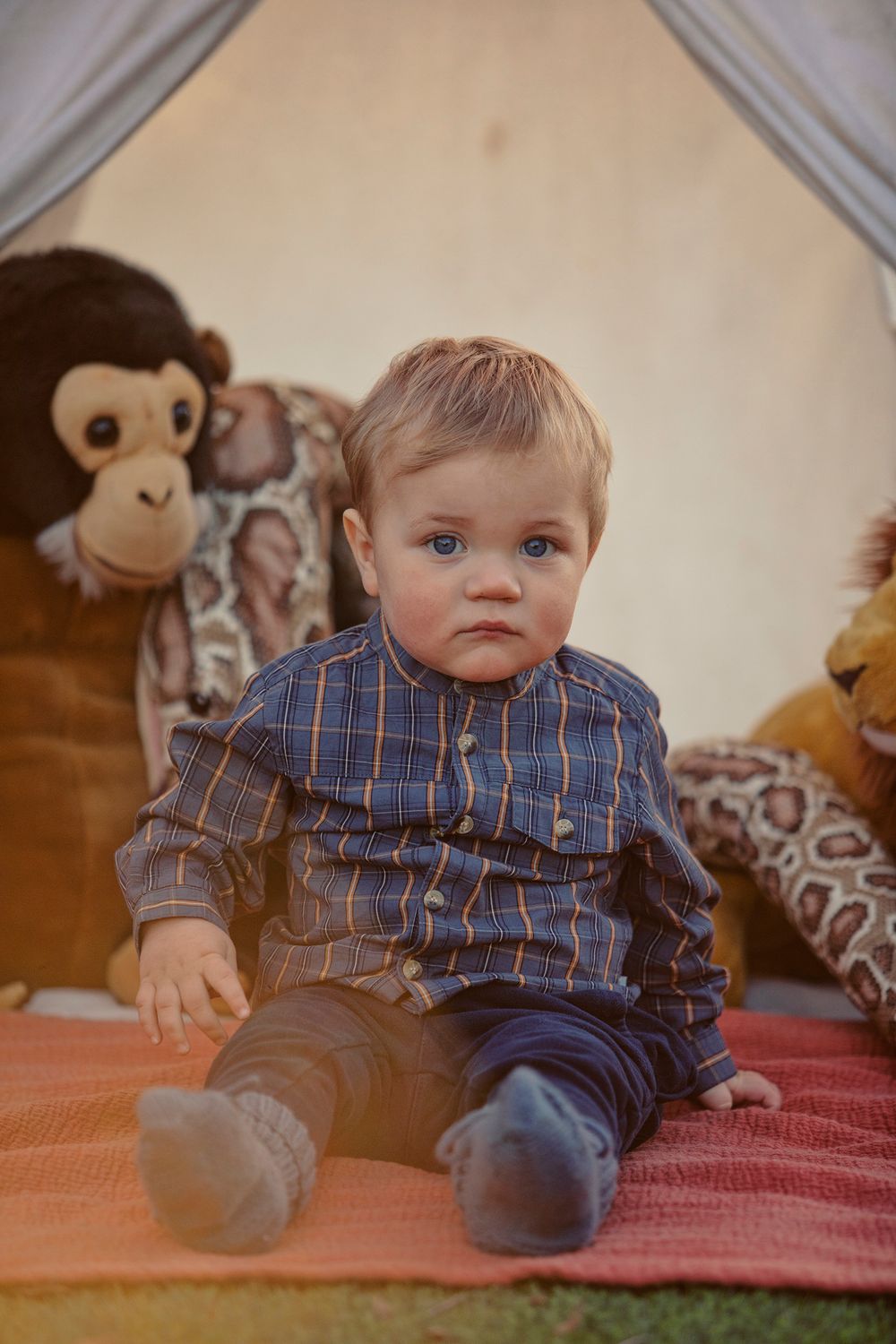 Cute baby boy in plaid shirt sits on orange blanket surrounded by stuffed animal toys in a teepee tent.