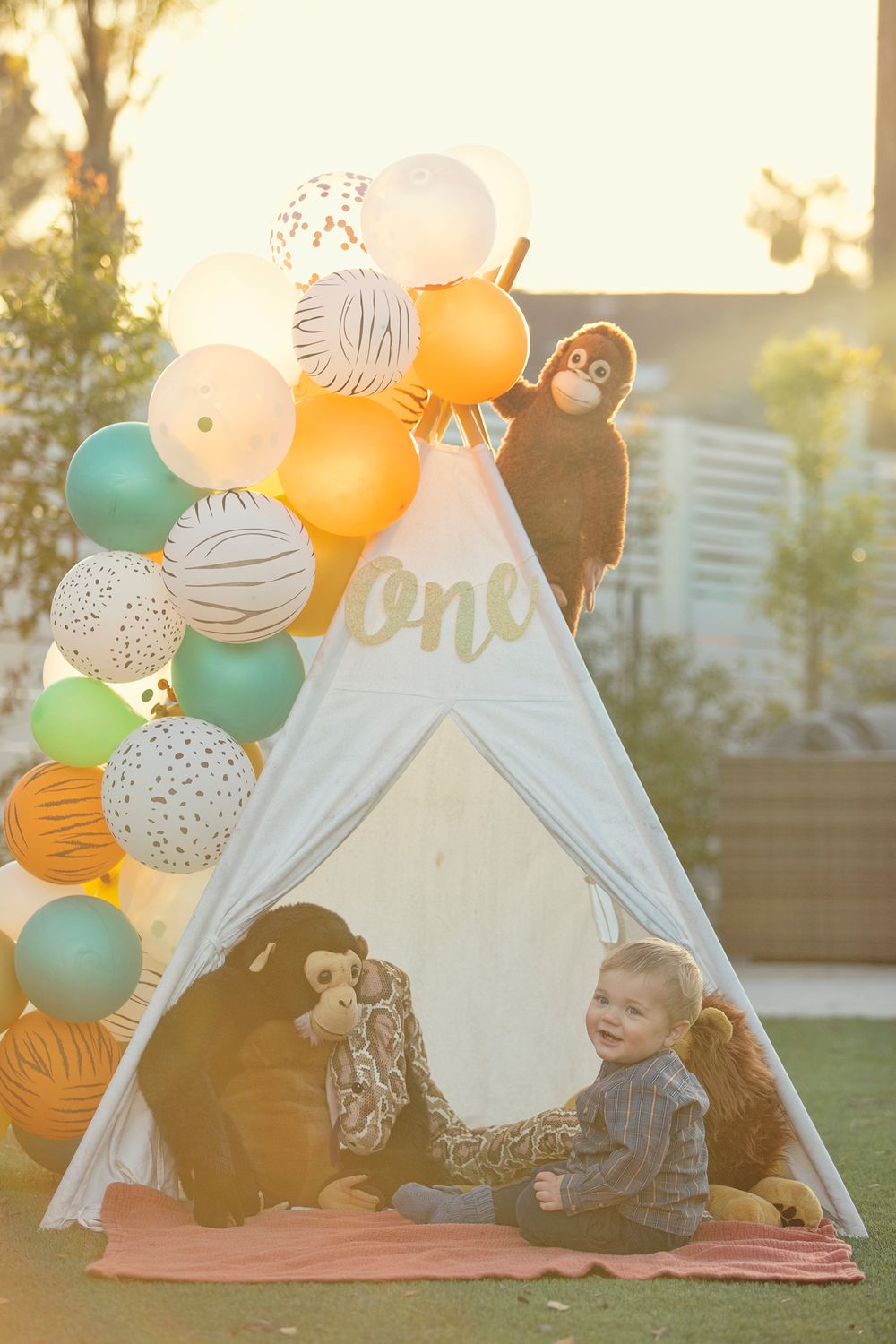 Toddler boy sitting inside a white teepee with stuffed animals and colorful safari-themed balloons at a first birthday party.