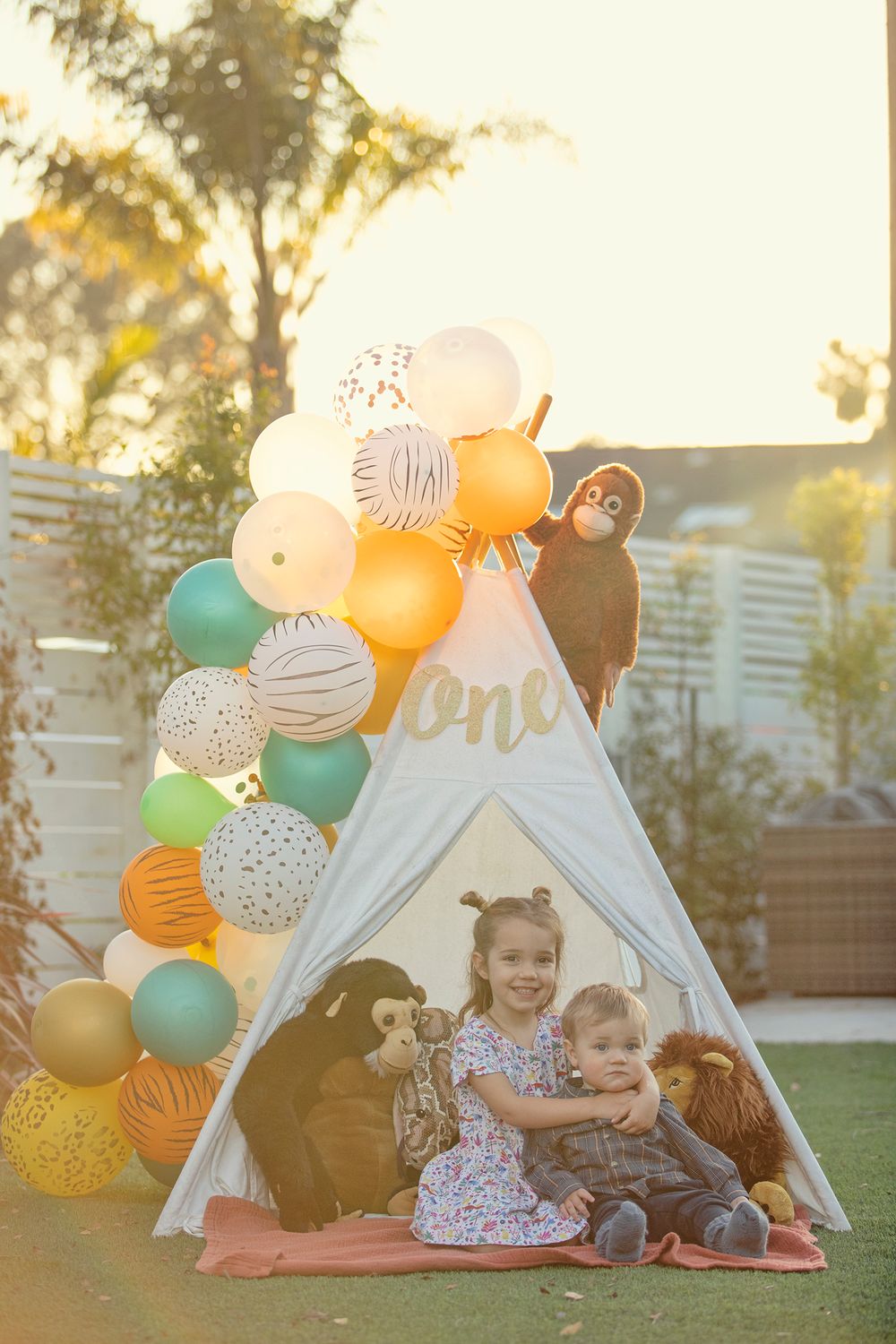Two toddlers sit inside a teepee surrounded by stuffed animals and colorful safari-themed balloons at a first birthday party.