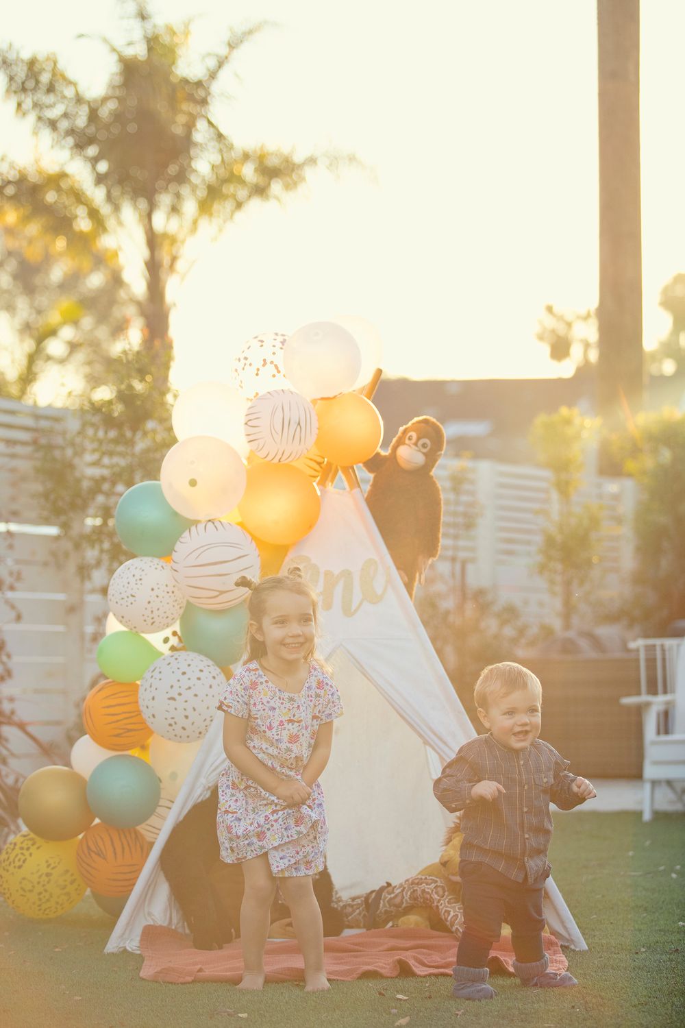 Two young children standing in front of a white teepee decorated with colorful safari balloons at a first birthday party.