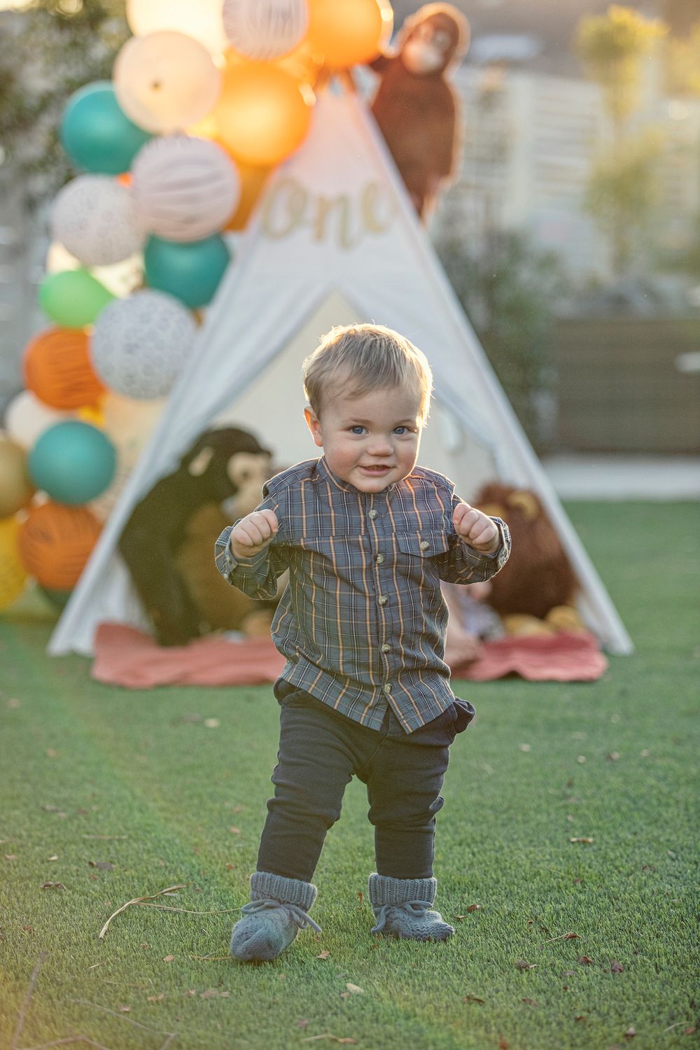 Adorable toddler boy in plaid shirt stands on grass in front of a teepee with colorful balloons at first birthday party.