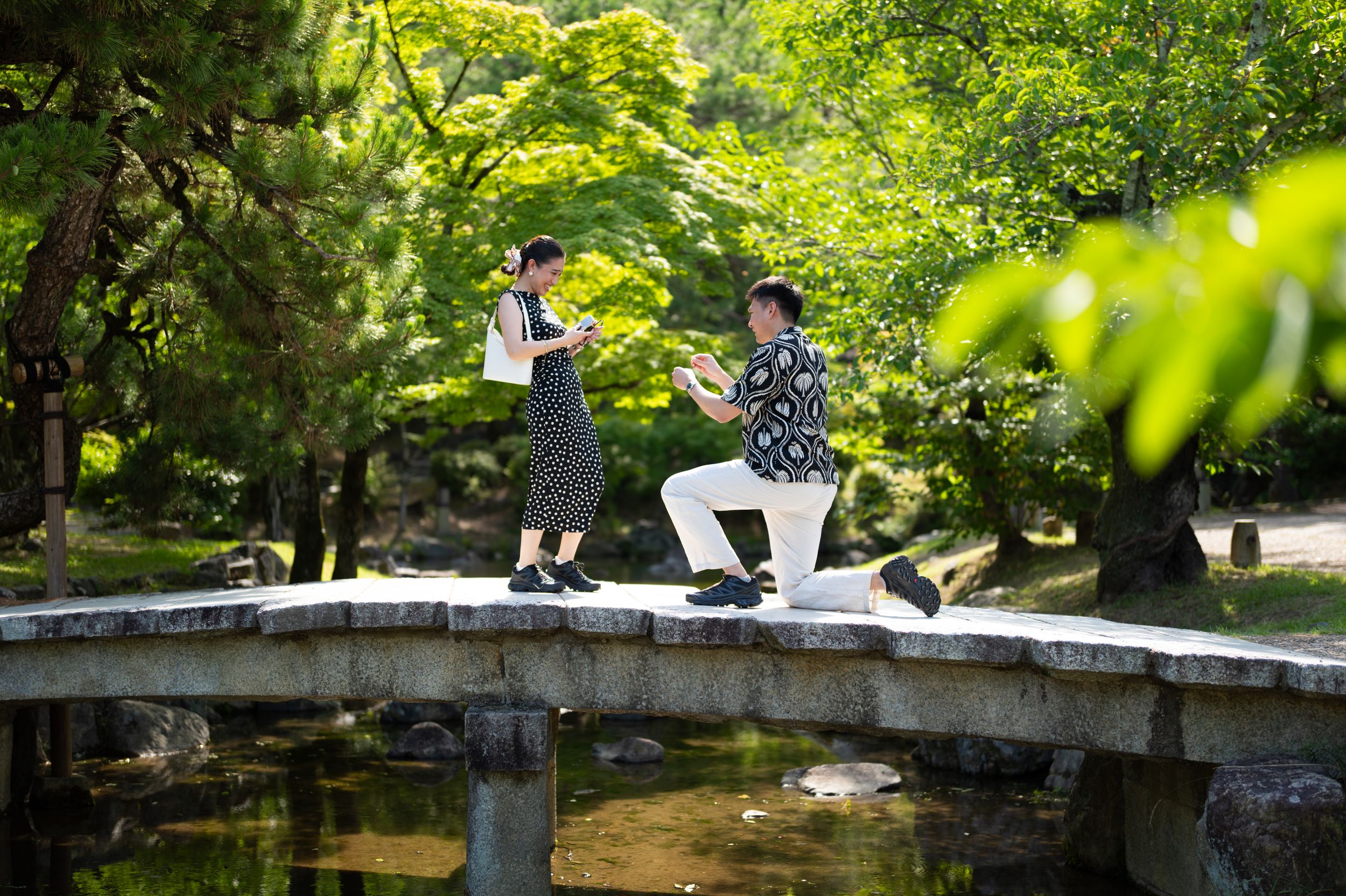 Couple standing on a wooden bridge surrounded by lush green trees in a Japanese garden.