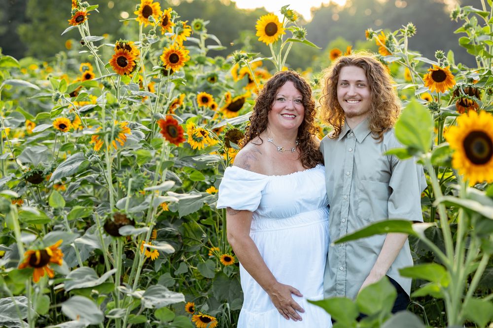 Couple smiling in a vibrant sunflower field, woman in white dress, man in green shirt, golden hour lighting.