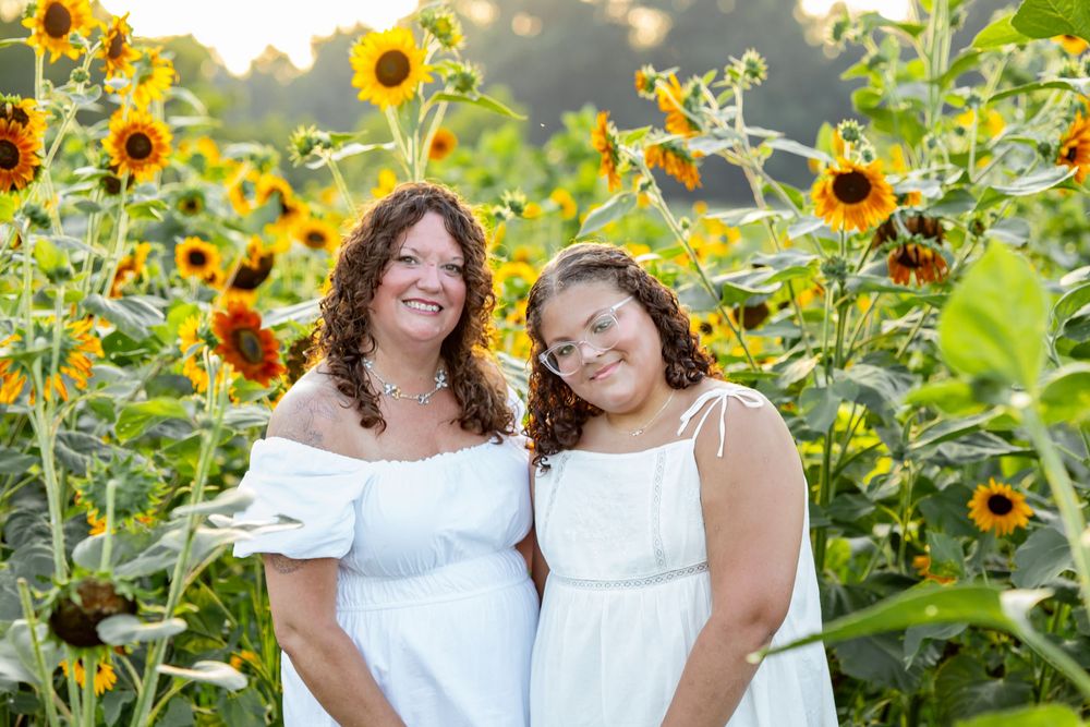 Two women in white dresses smile together in a vibrant sunflower field during golden hour.