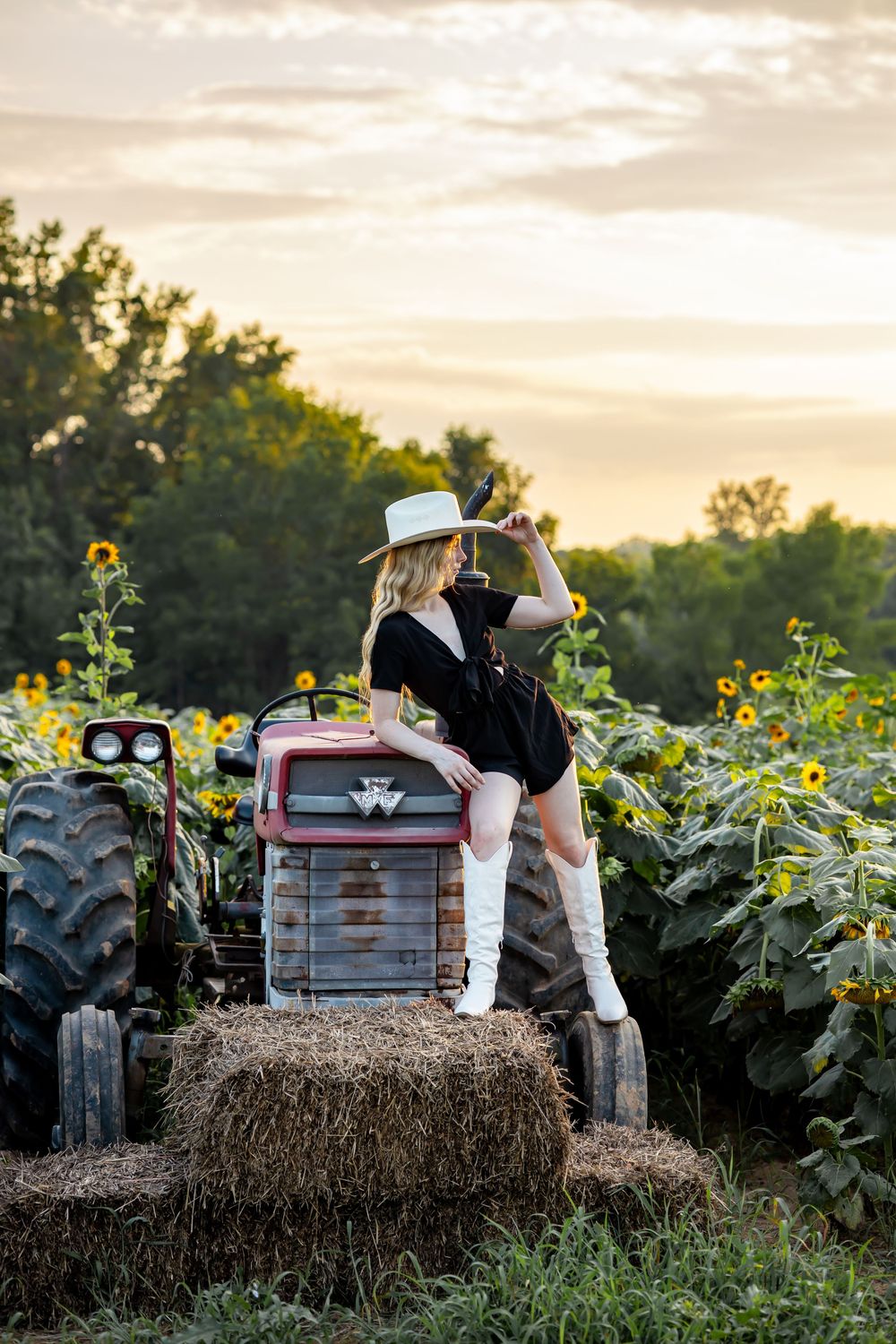 Woman in black dress, white boots, and cowboy hat poses on a tractor in a sunflower field at sunset.