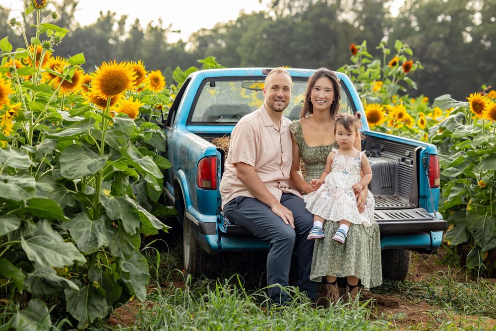 Family of three sitting on a teal pickup truck tailgate surrounded by sunflowers in a sunny field.