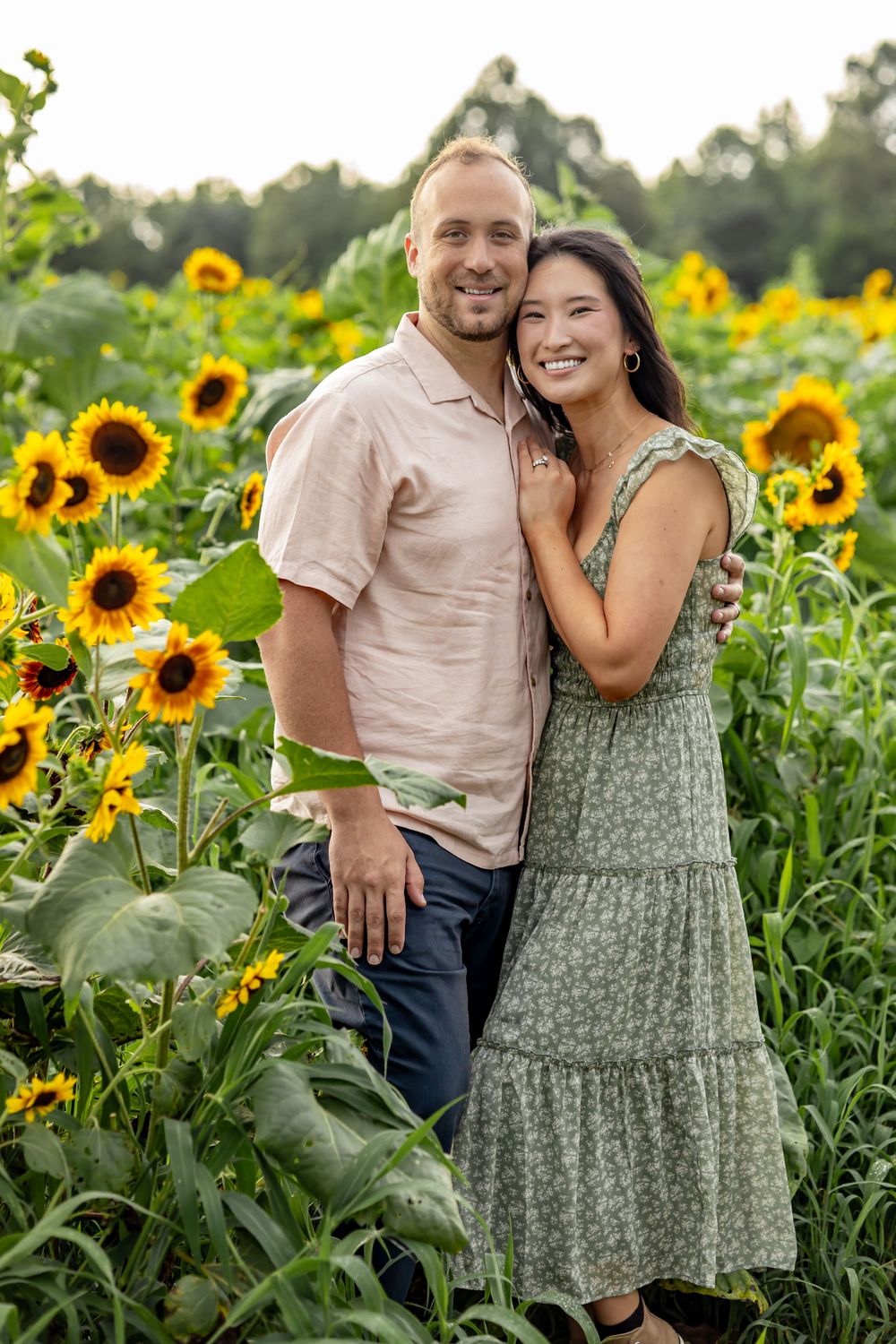 A smiling couple embraces in a vibrant sunflower field, woman in green floral dress, man in pink shirt, lush trees behind.