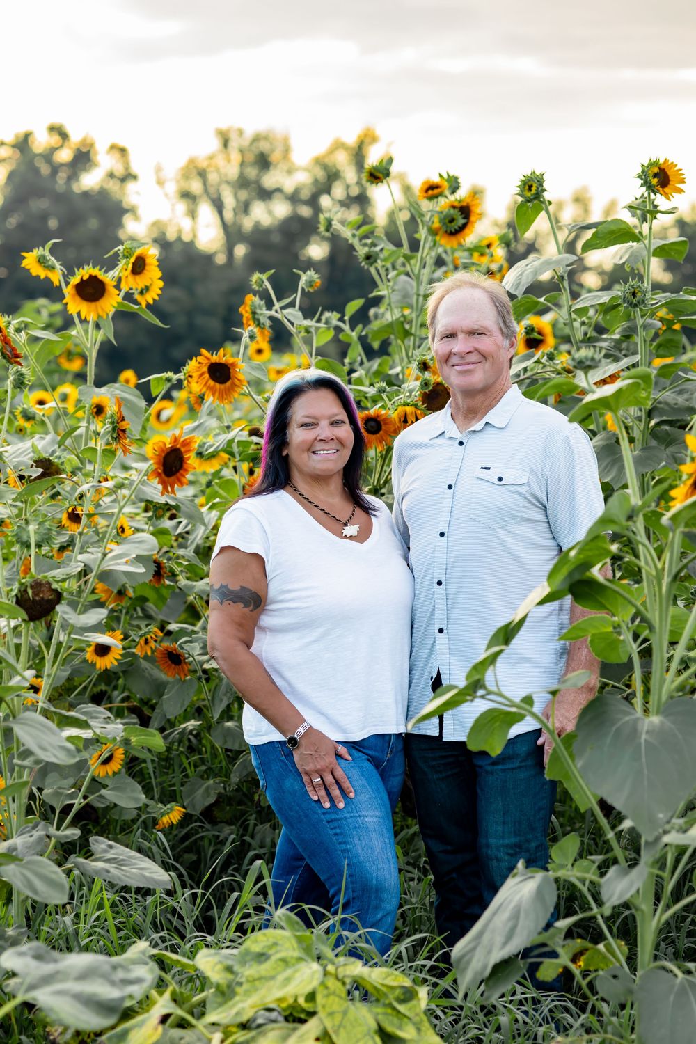 A smiling couple poses together in a vibrant sunflower field during golden hour sunset.