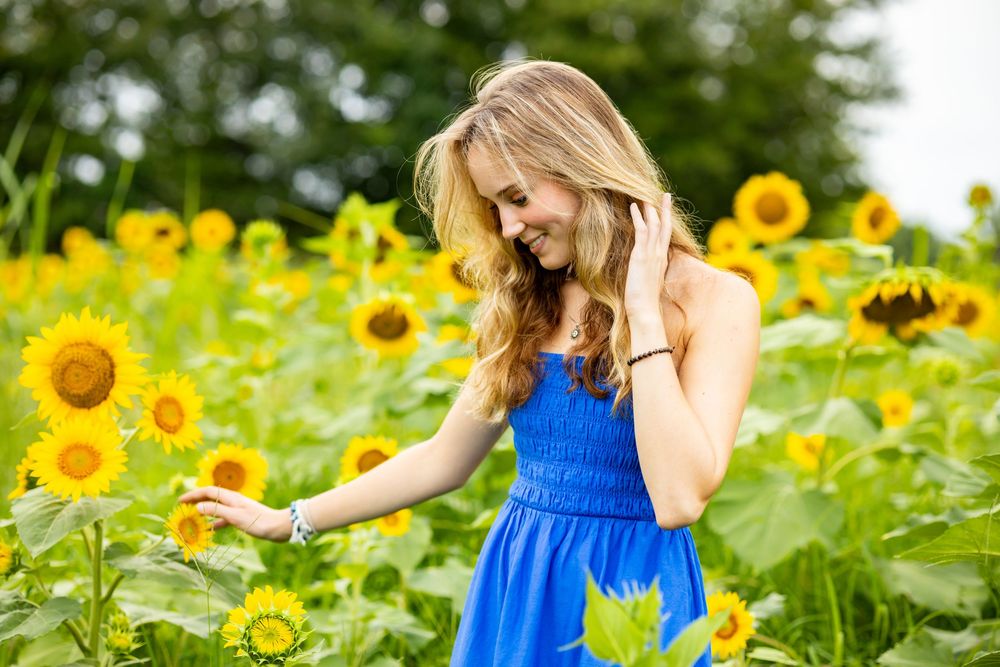 Young blonde woman in blue dress smiling among vibrant sunflowers in a sunny field.