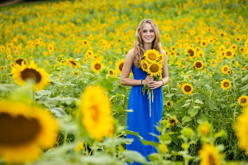 A smiling woman in a blue dress holds sunflowers while standing in a vibrant yellow sunflower field.