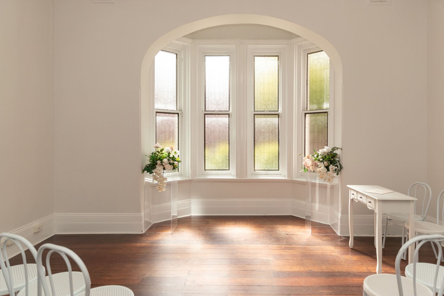 Bright wedding ceremony room with arched bay windows, white chairs, hardwood floors, and floral arrangements in natural light.