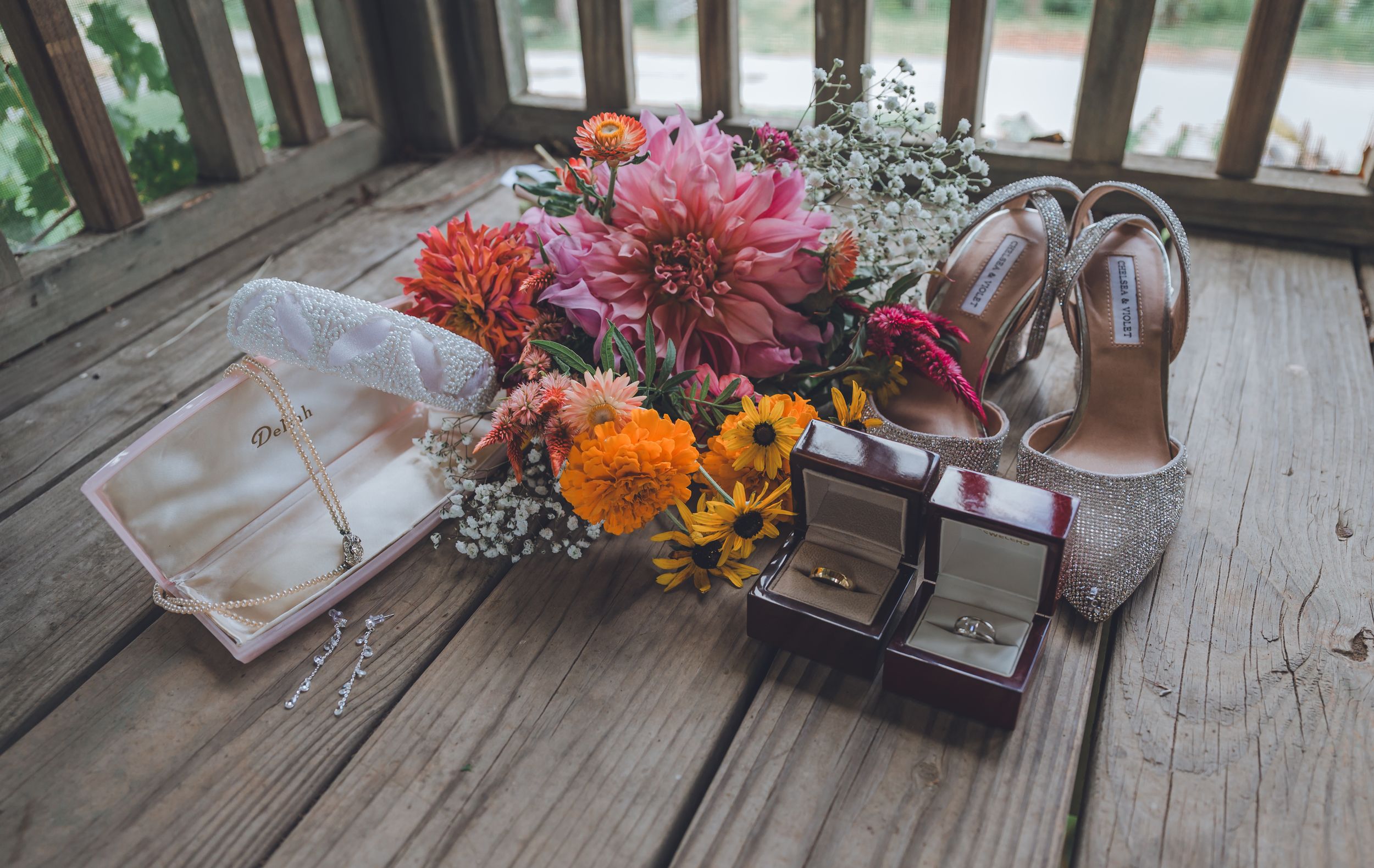 Wedding flat lay with colorful floral bouquet, glitter heels, rings, and jewelry on rustic wood deck.