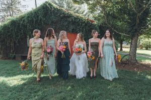 Bride and bridesmaids in mismatched dresses walk together outdoors near ivy-covered barn at rustic wedding.