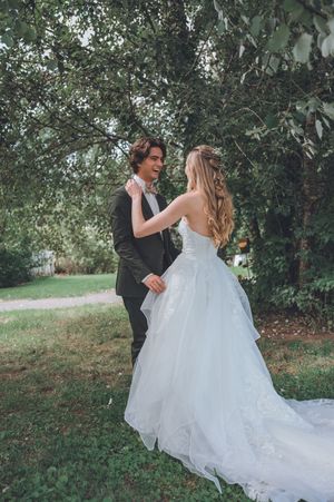 Bride in white ballgown and groom in gray suit share a romantic moment outdoors under green trees.