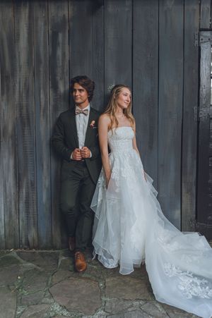Bride in strapless lace gown and groom in dark suit with bow tie lean against rustic wooden barn wall.