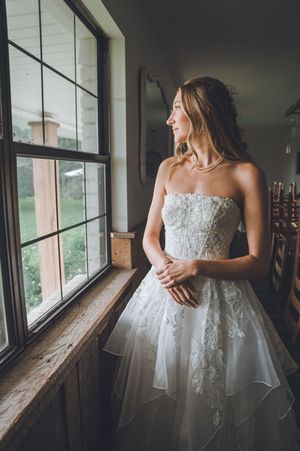 Bride in strapless lace wedding gown gazing out a rustic window, smiling softly in natural light.