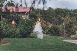 Bride in white layered gown walks through lush garden with colorful flowers at outdoor wedding venue.