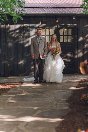 Bride in white gown walks arm-in-arm with escort on stone path outside rustic dark barn venue.