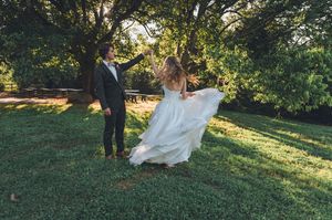 Groom spinning bride in flowing white wedding dress outdoors under large trees on lush green grass.