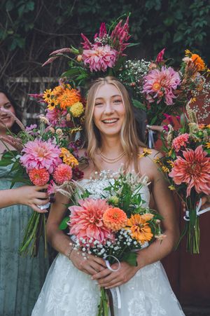 Smiling bride holding colorful dahlia bouquet with large floral crown of pink and orange blooms at outdoor wedding.