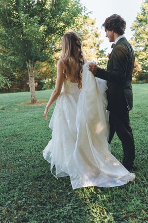 Bride in white lace gown and groom in black suit walk hand in hand across green lawn with trees.