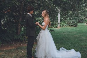 Bride in white gown and groom in dark suit exchange vows outdoors in lush green garden setting.