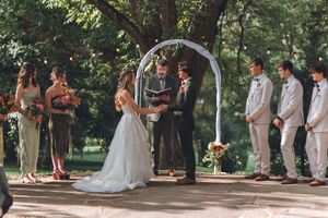 Outdoor wedding ceremony under floral arch with bride, groom, officiant, bridesmaids, and groomsmen in lush garden setting.