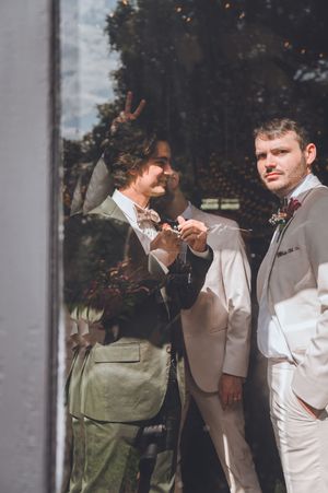 Two grooms in elegant suits standing by a glass window at their wedding ceremony.